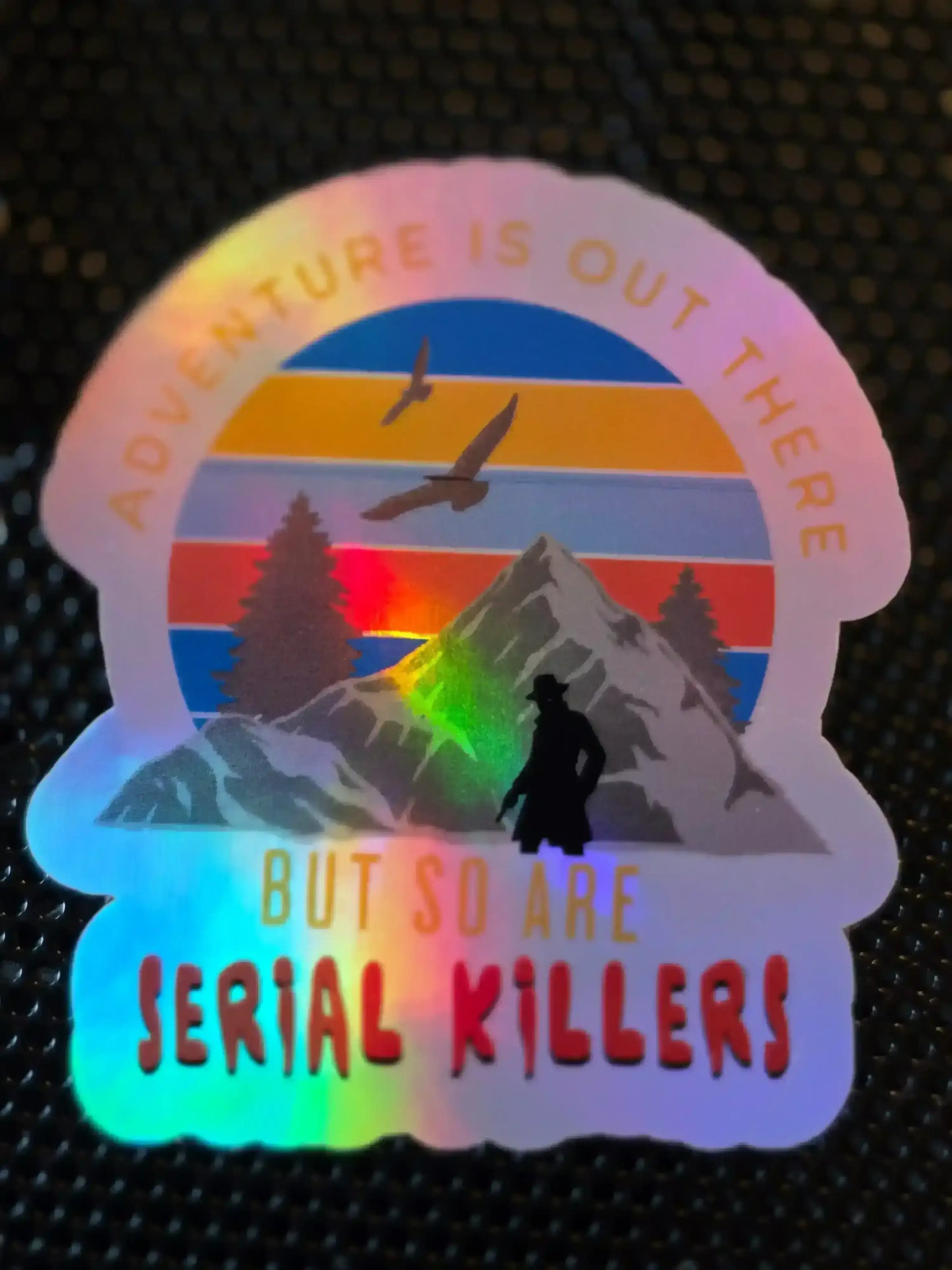 Holographic sticker with mountain, birds, and silhouette, reads 'Adventure is out there but so are serial killers'