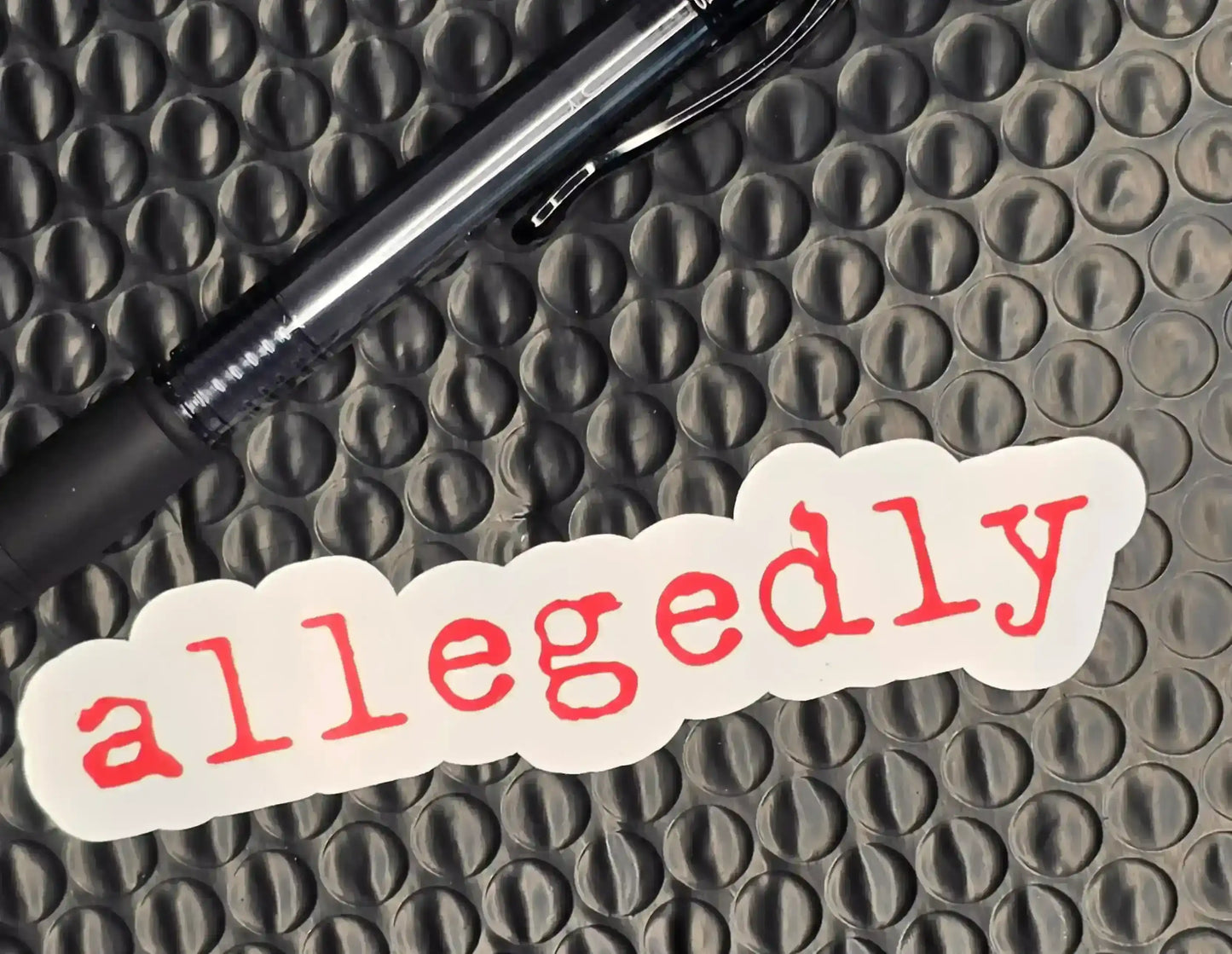 Allegedly sticker with red text on white, next to black pen on textured surface, Cady Creations