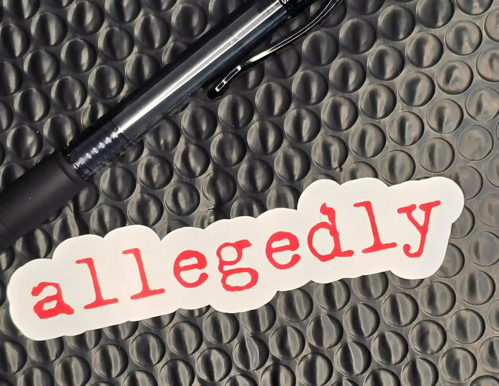 Allegedly sticker with red text on white, next to black pen on textured surface, Cady Creations