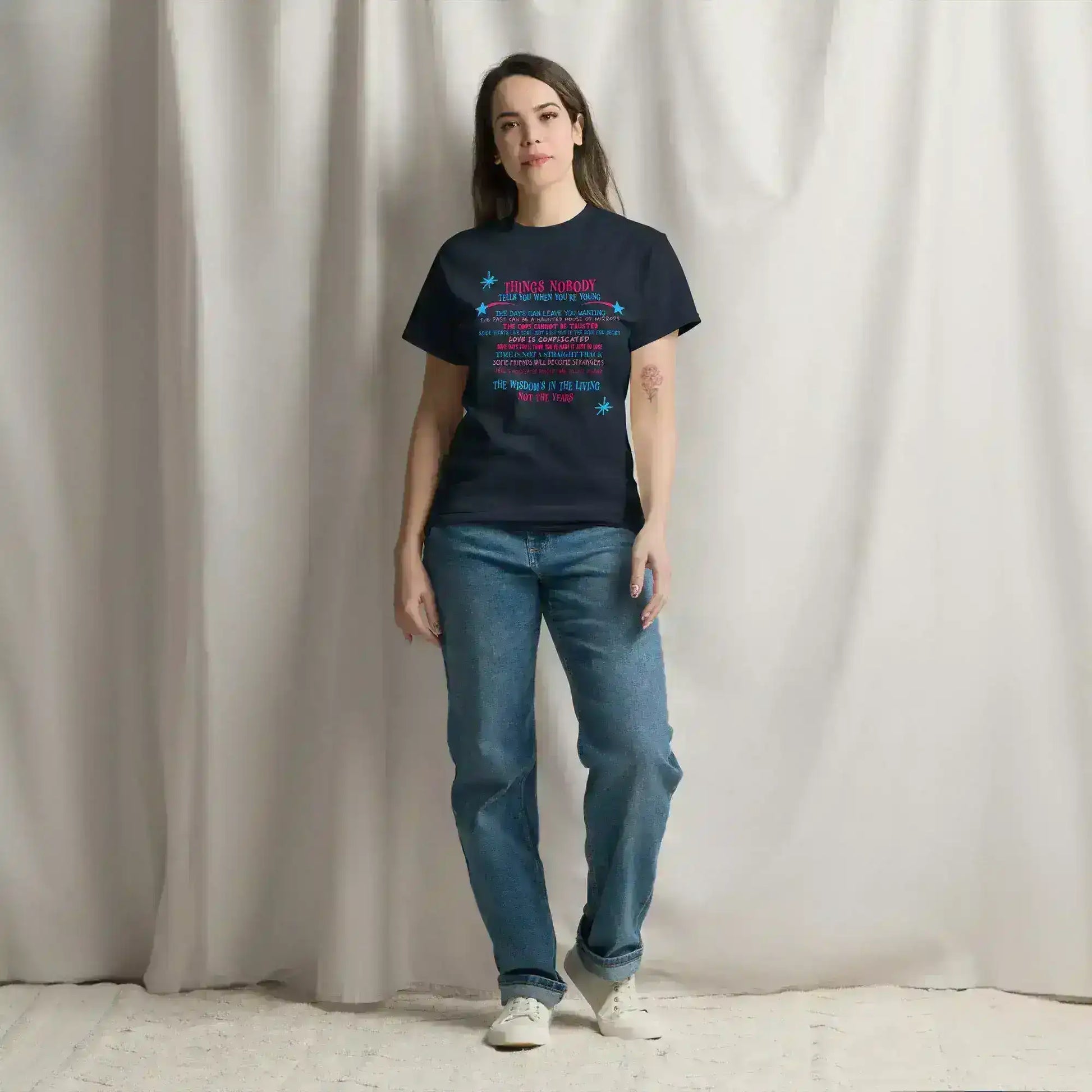 Woman wearing Cady Creations navy graphic tee and jeans, standing indoors against a curtain.